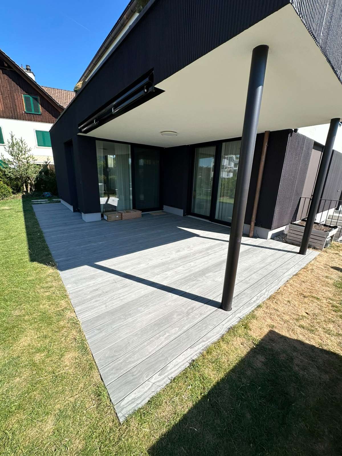 Moderne Accoya Terrasse aus Accoya Grey mit grauen Terrassendielen vor einem schwarzen Einfamilienhaus und überdachtem Sitzbereich.